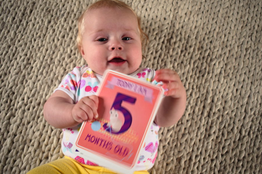 Baby lying on a rug, holding a card which says 'Today I am five months old'