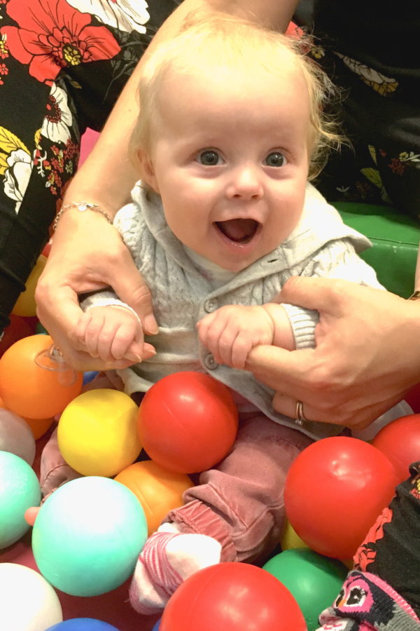 Baby sitting in ball pit with excited expression, holding onto the hands of the mother, who is sitting behind her