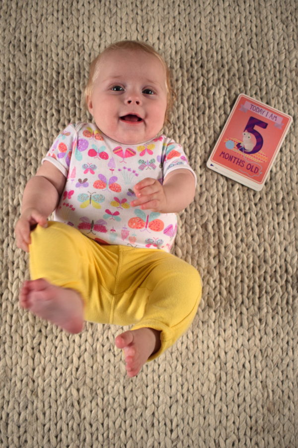 Baby lying on her back with her feet in the air with a card next to her saying 'I am five months old'