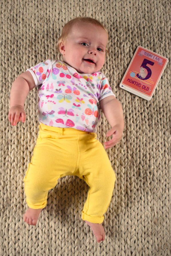 Baby in yellow trousers, lying on the floor with a card next to her which reads 'Today I am five months old'