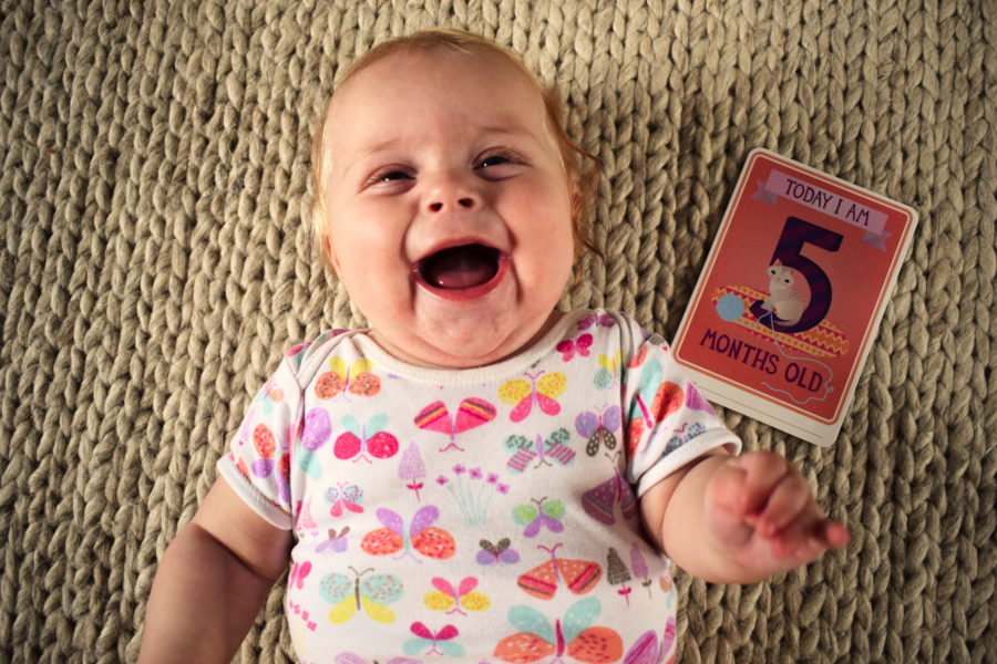 Baby lying on her back, open mouthed laughing, with a card next to her saying 'Today I am five months old'
