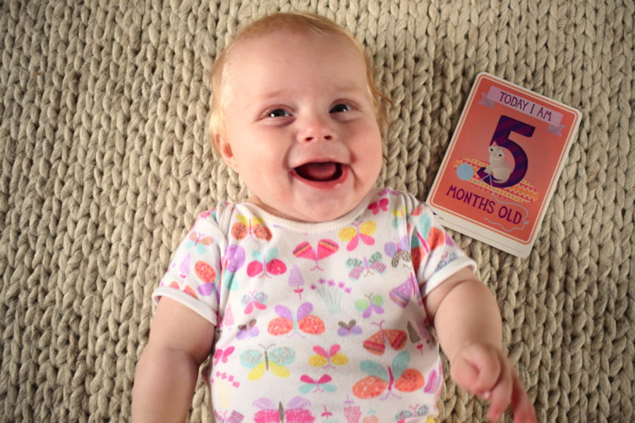 Baby lying on her back, smiling, with a card next to her, which reads, 'today I am five months old'