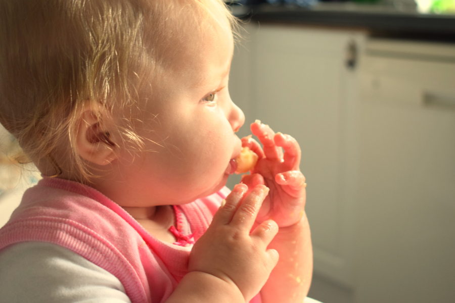 Side profile of baby covered in banana in  pink bib, eating a piece of banana