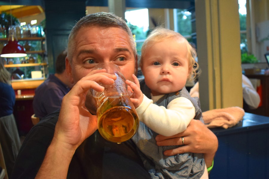 father holding baby girl, drinking from a pint of beer, with baby holding on to the glass