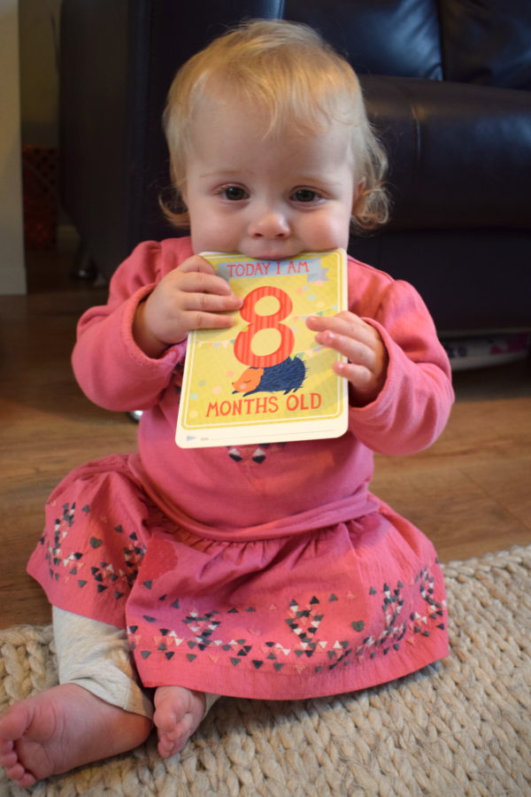 baby girl in pink top sitting up and biting a milestone card saying today I am eight months old
