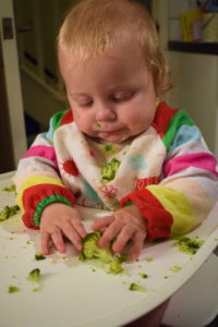 baby in highchair wearing bib, holding broccoli