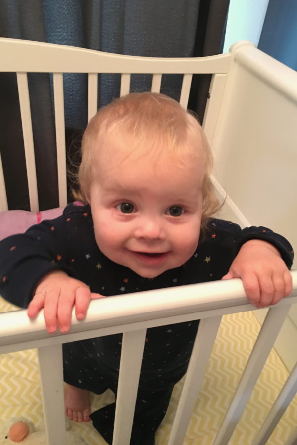 nine month old girl standing up in cot, in blue pyjamas, smiling