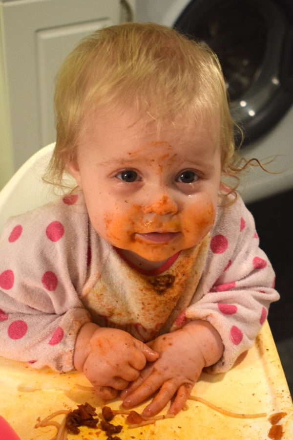 baby in highchair, covered in tomato sauce with spaghetti in front of her