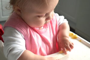 baby sitting in highchair, picking up a piece of banana, wearing pink bib