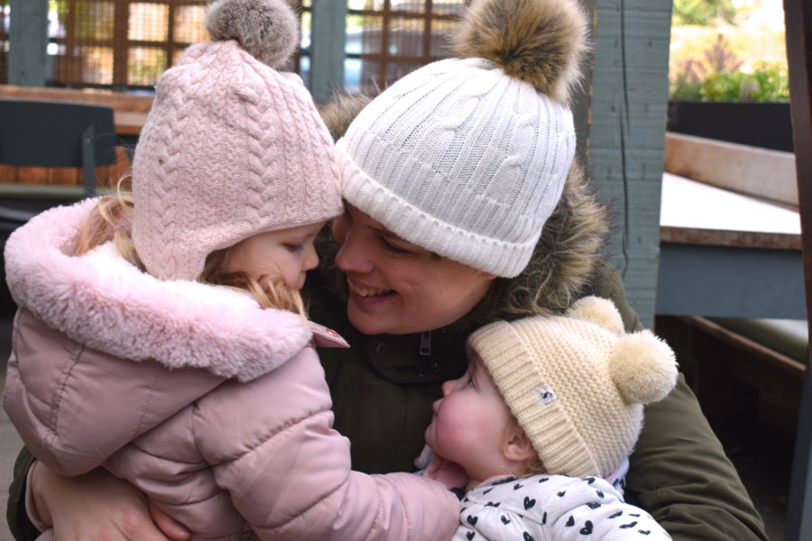 A mother holding two little girls, all looking at each other and smiling, each wearing a bobble hat and coat