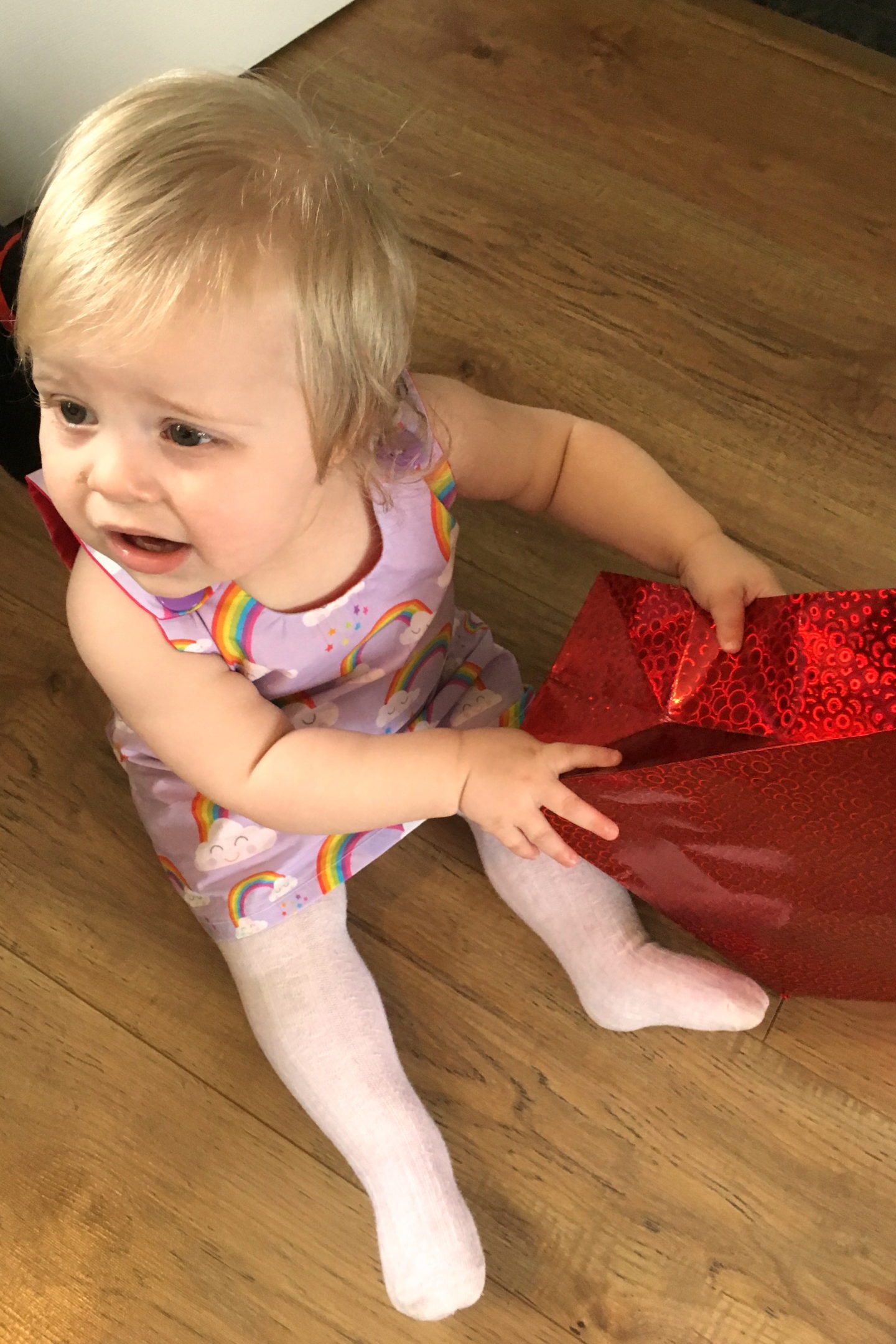 one year old in party dress sitting on the floor holding red gift bag