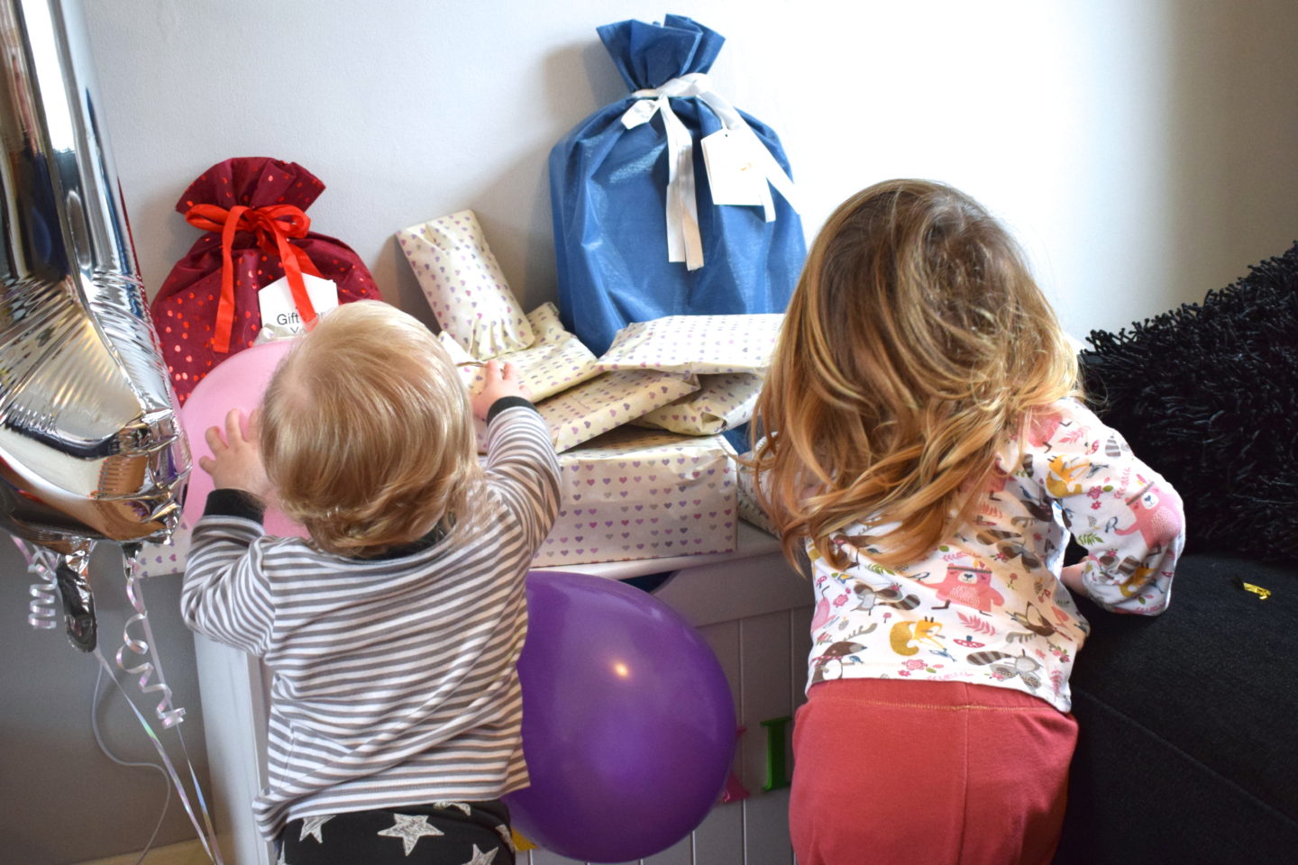 one year old girls with older sister with back to camera, looking at pile of birthday presents