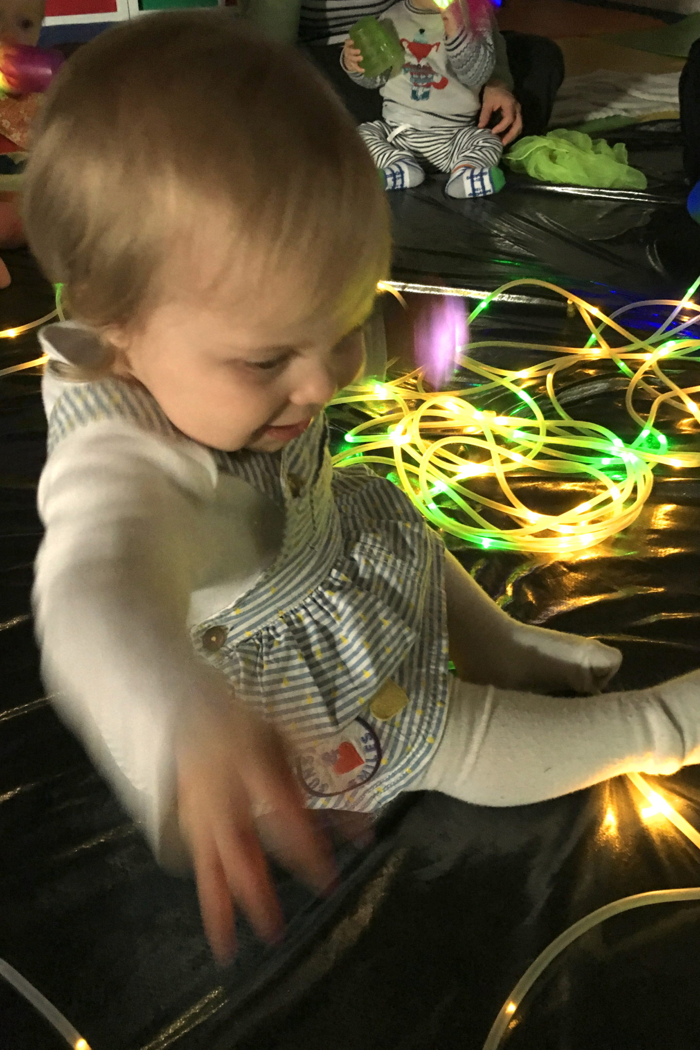 one year old girl in blue dress, playing with string lights, sitting on the floor