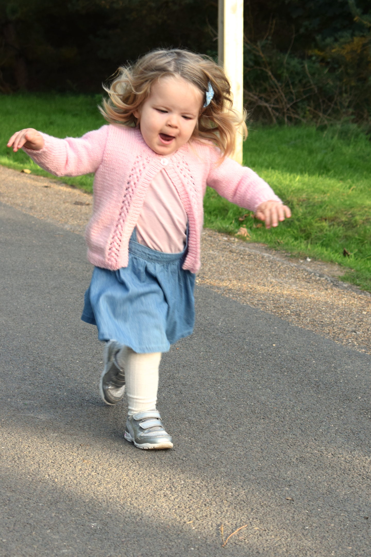 two and a half year old girl running down a path with arms outstretched, smiling, in pink cardigan and blue dress