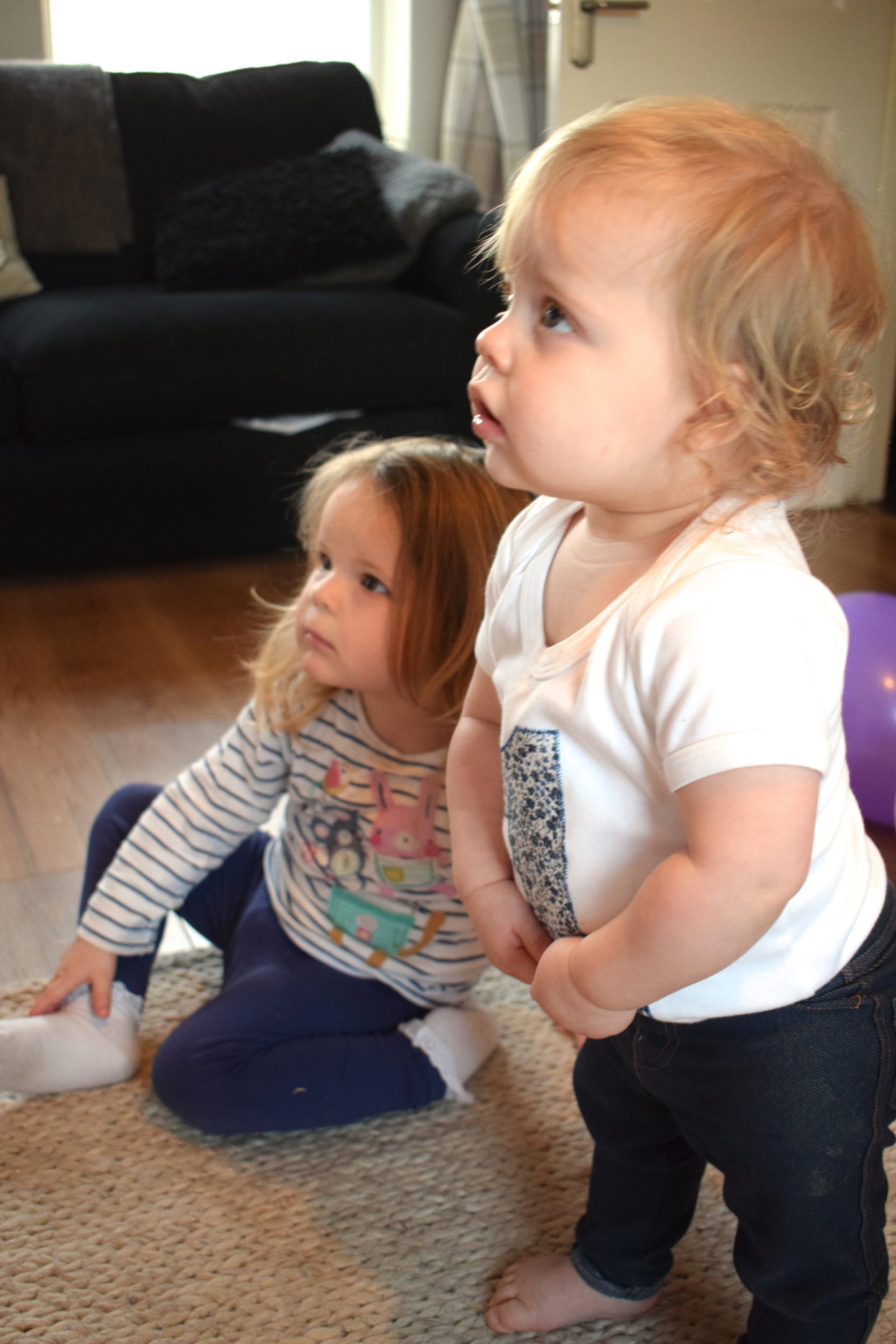 sisters, one standing, one sitting on the floor, watching TV