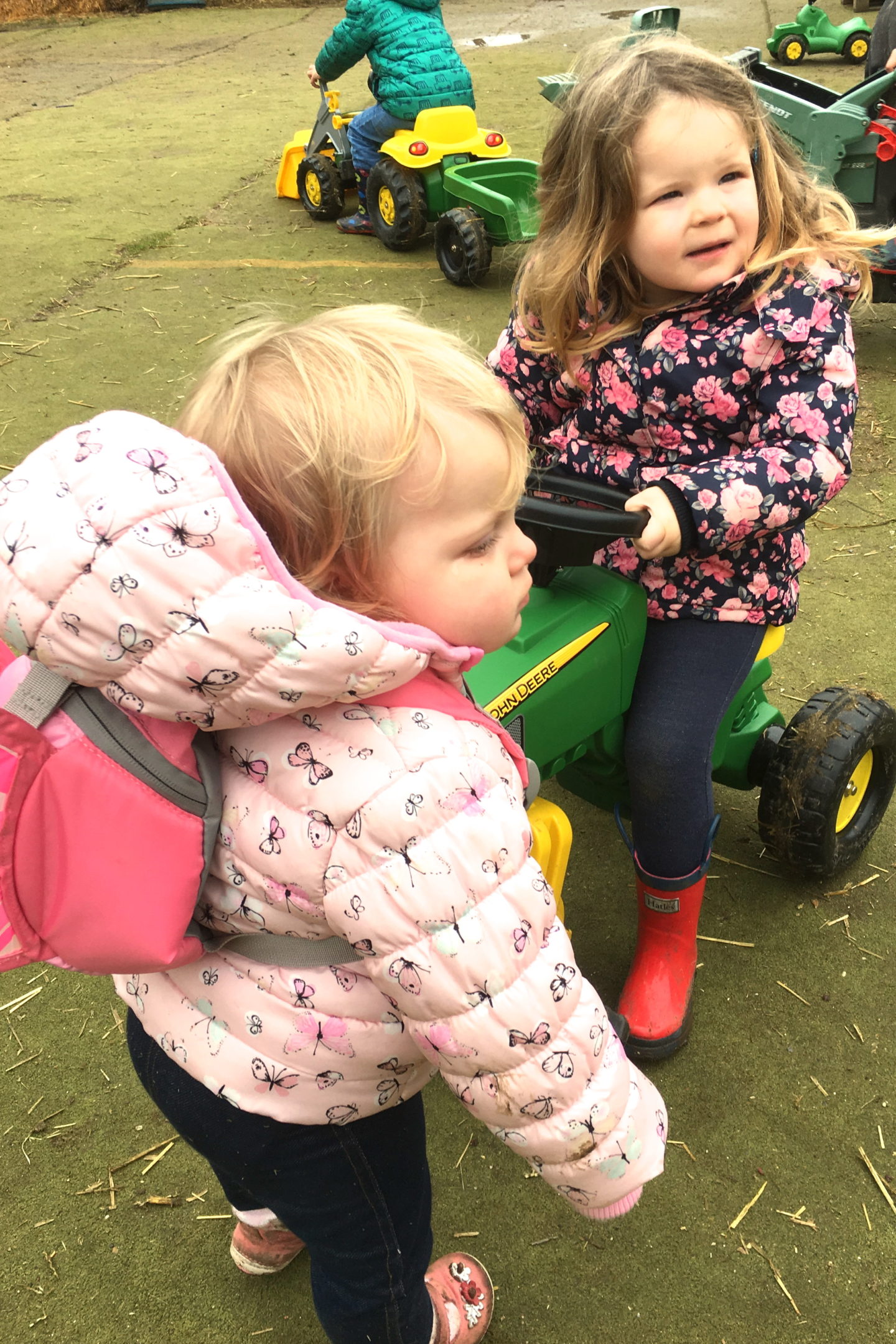 toddler sisters, one on toy tractor, the other looking on