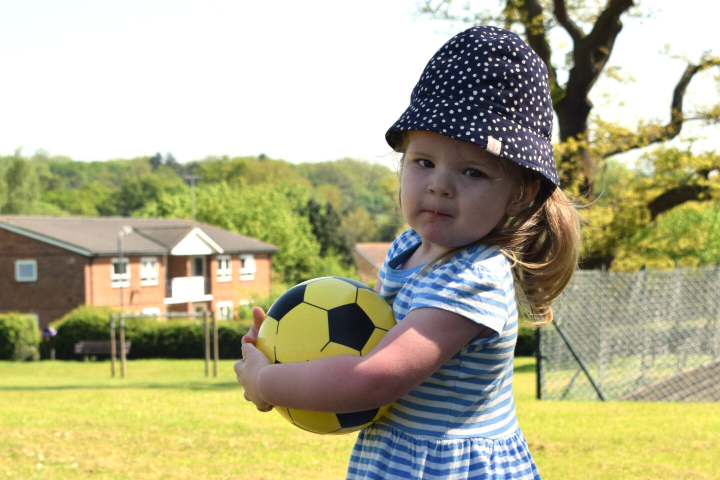 Two year old girl who's recently had genetics testing in stripy dress and sunhat, holding ball, looking over her shoulder.