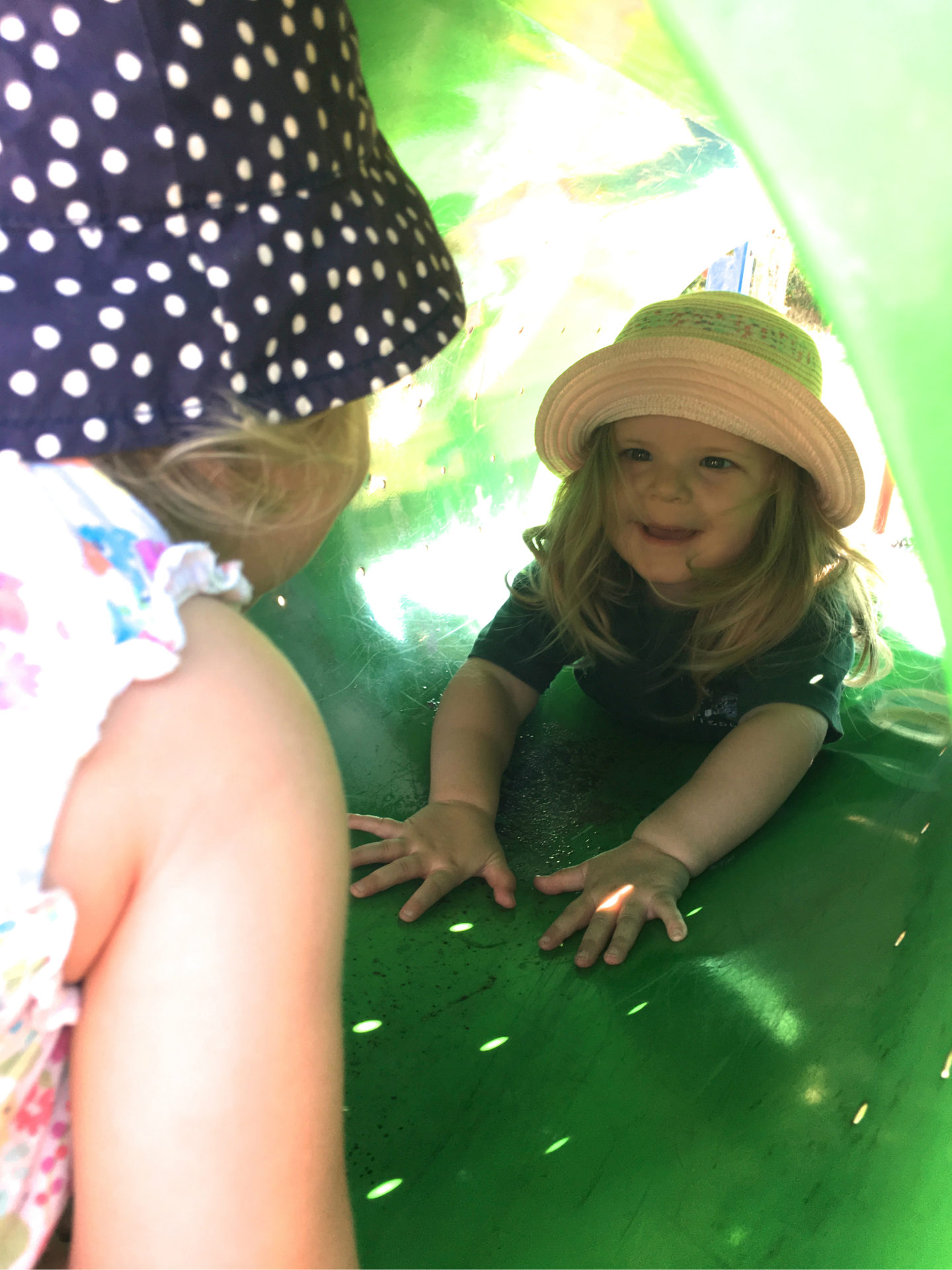 two year old in a tunnel at a playground, laughing at her sister who is looking into the tunnel
