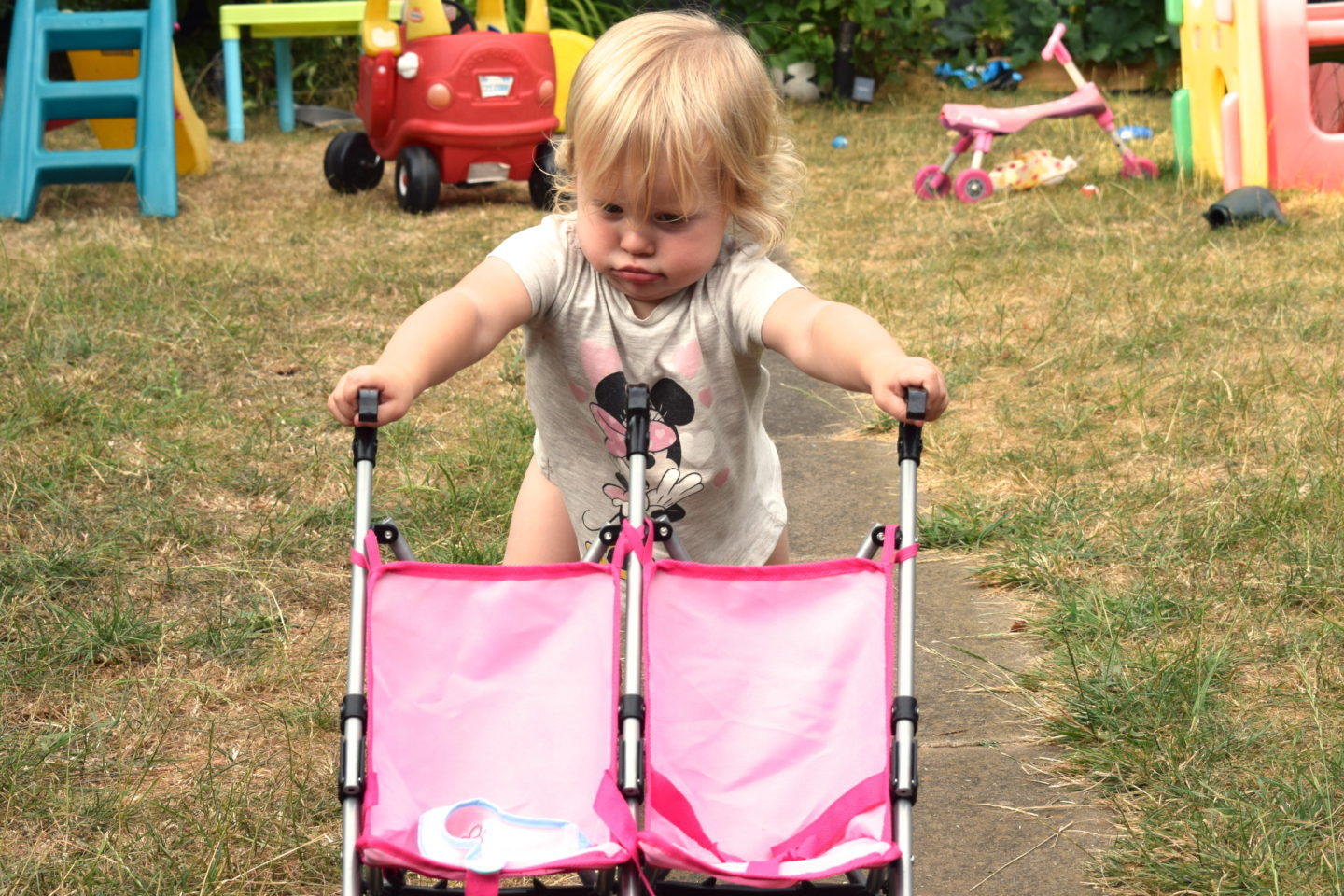 18 month old girl, pushing doll buggy up the garden path