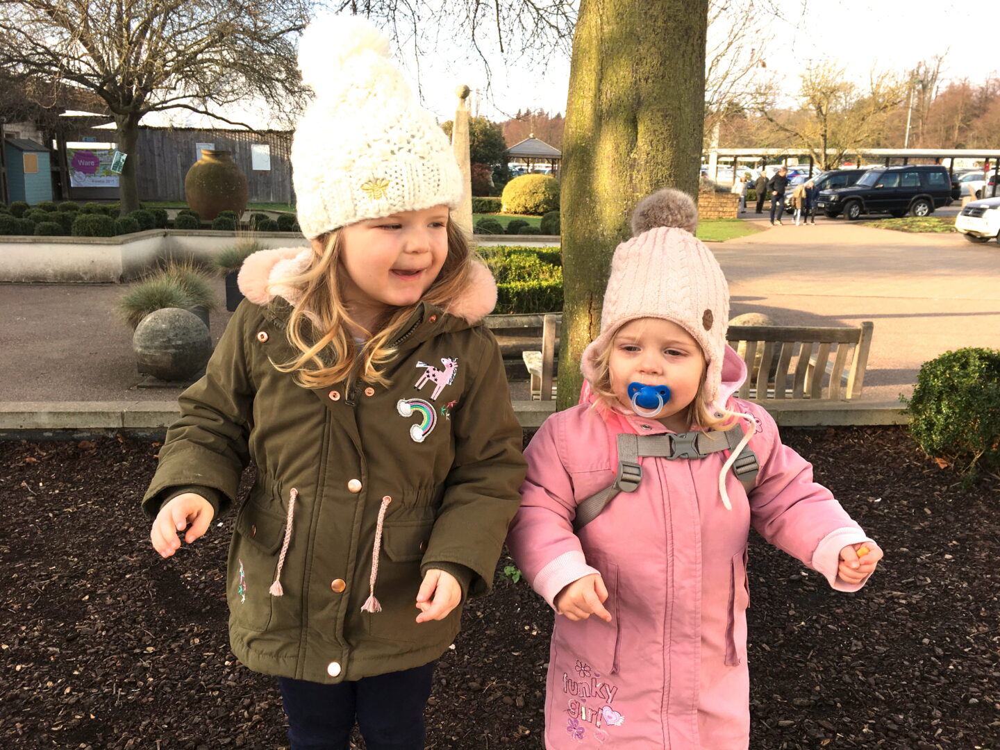 Two toddler sisters standing side by side on a wall in bobble hats and coats