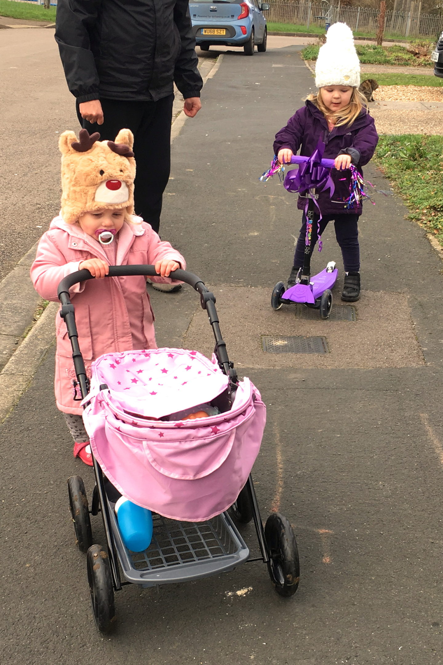 two sisters, one pushing a dolls pram, and one on a scooter, going out for a walk with their dad