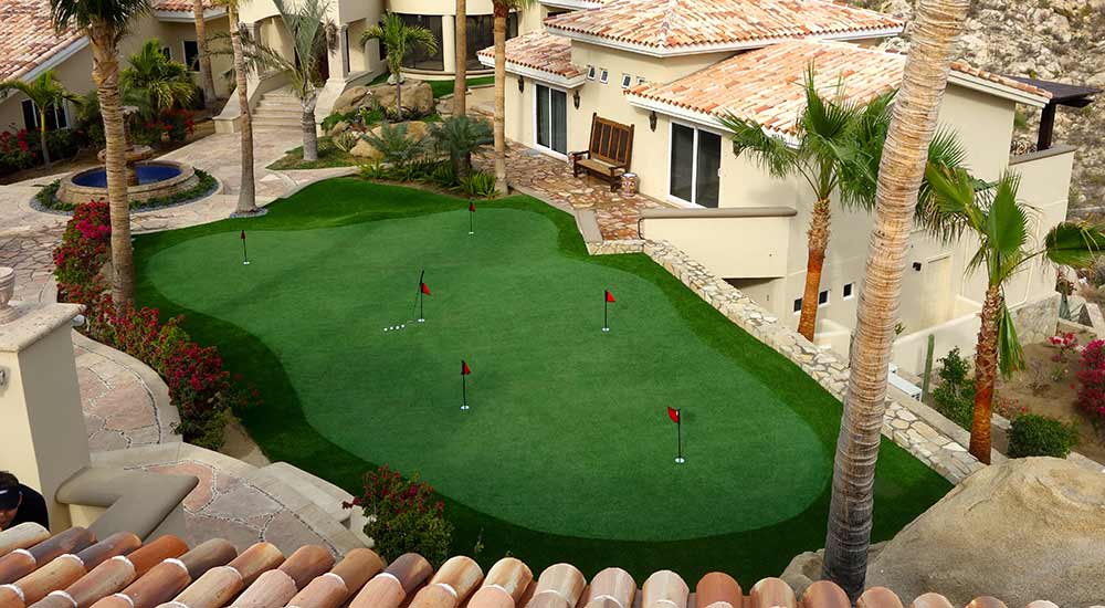 Golf green made from artificial grass, surrounded by palm trees and villas
