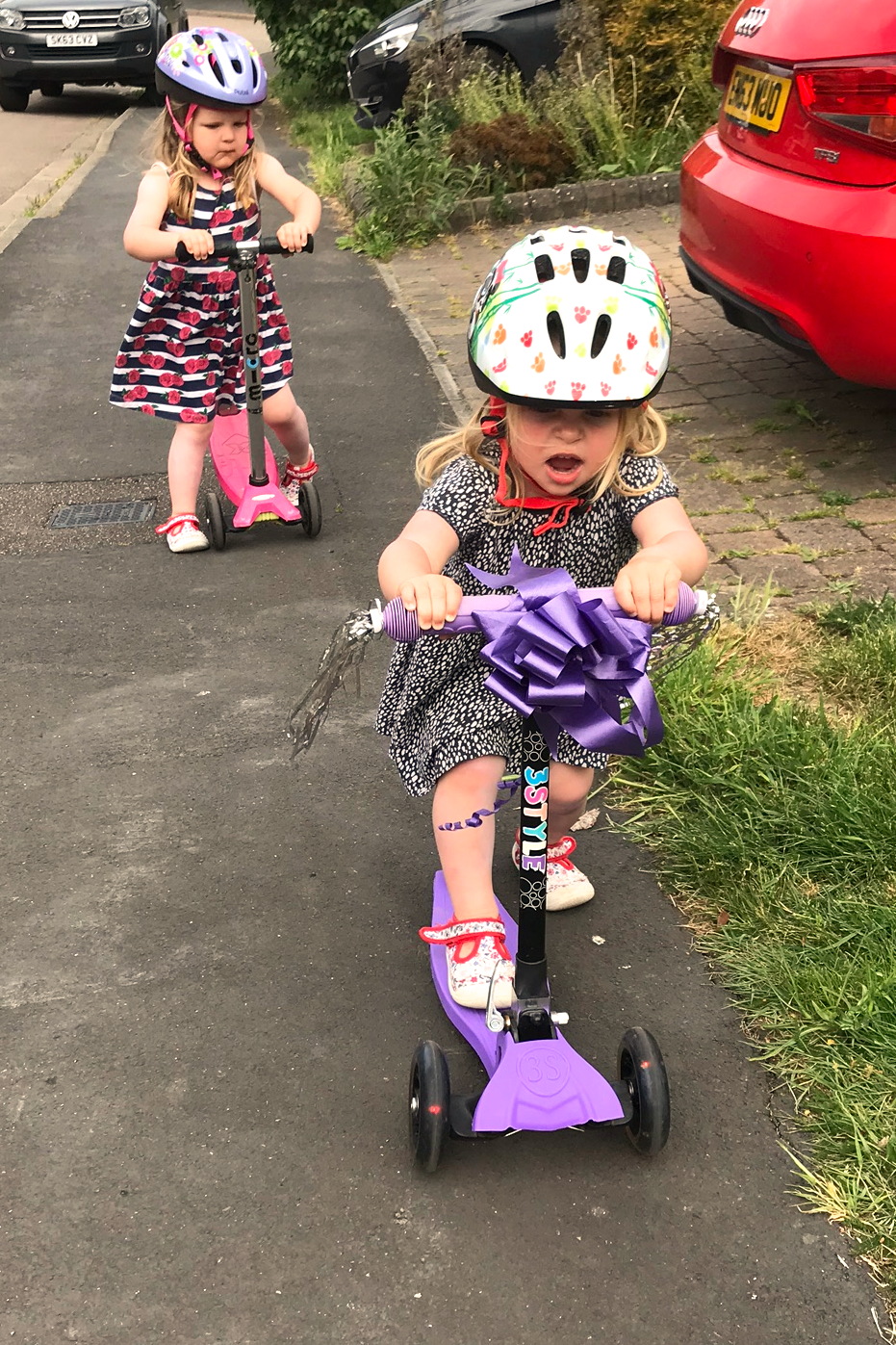 Two toddler sisters on scooters with helmets on, scooting down a pavement