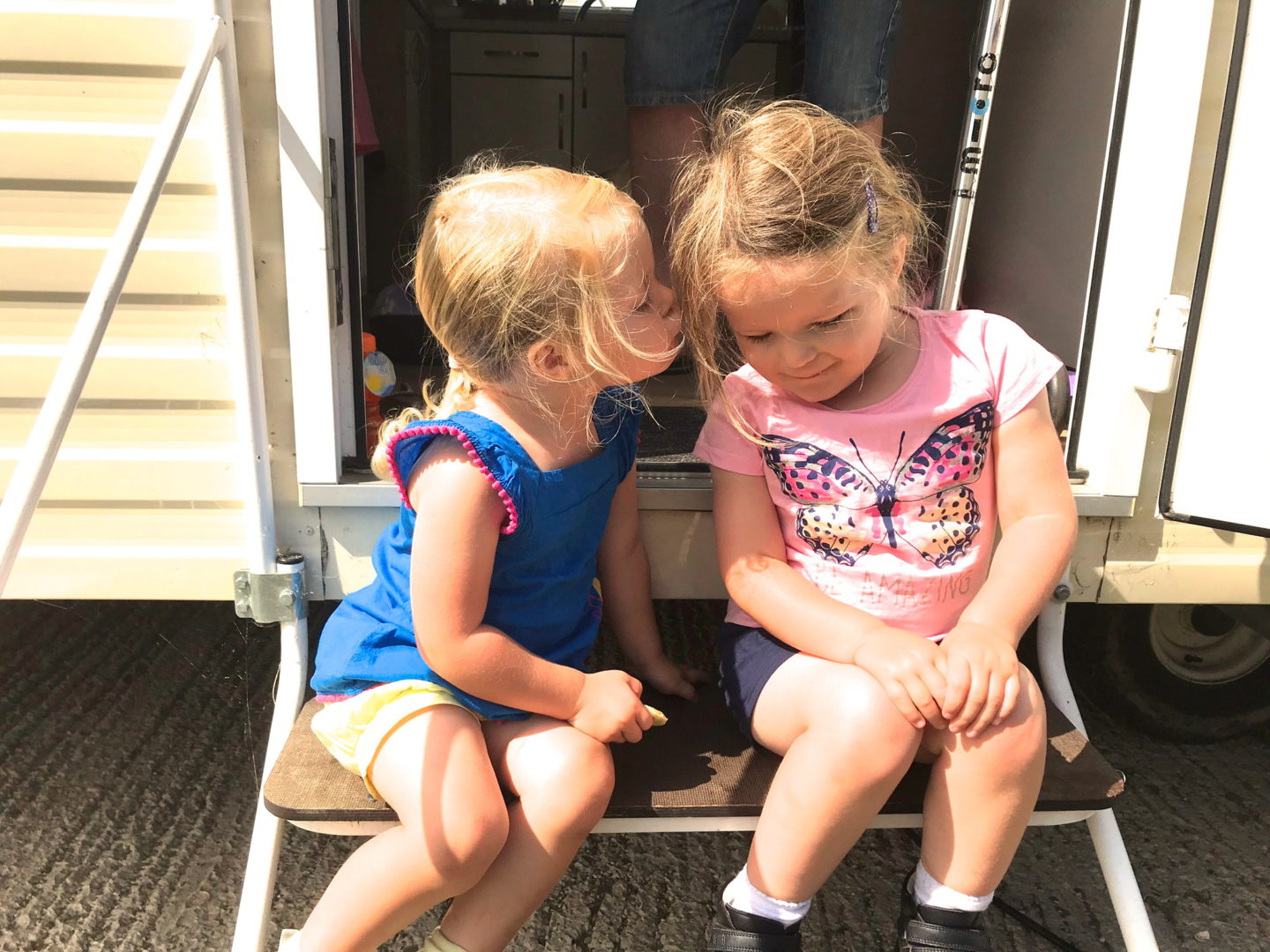 two little sisters sitting on the steps of a caravan, one kissing the other's cheek