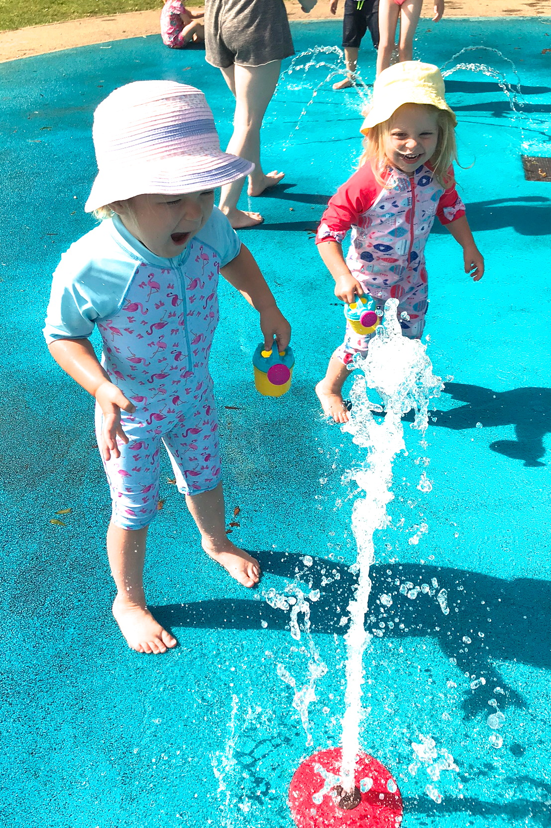 Two sisters in a splash park, playing with a jet of water, laughing